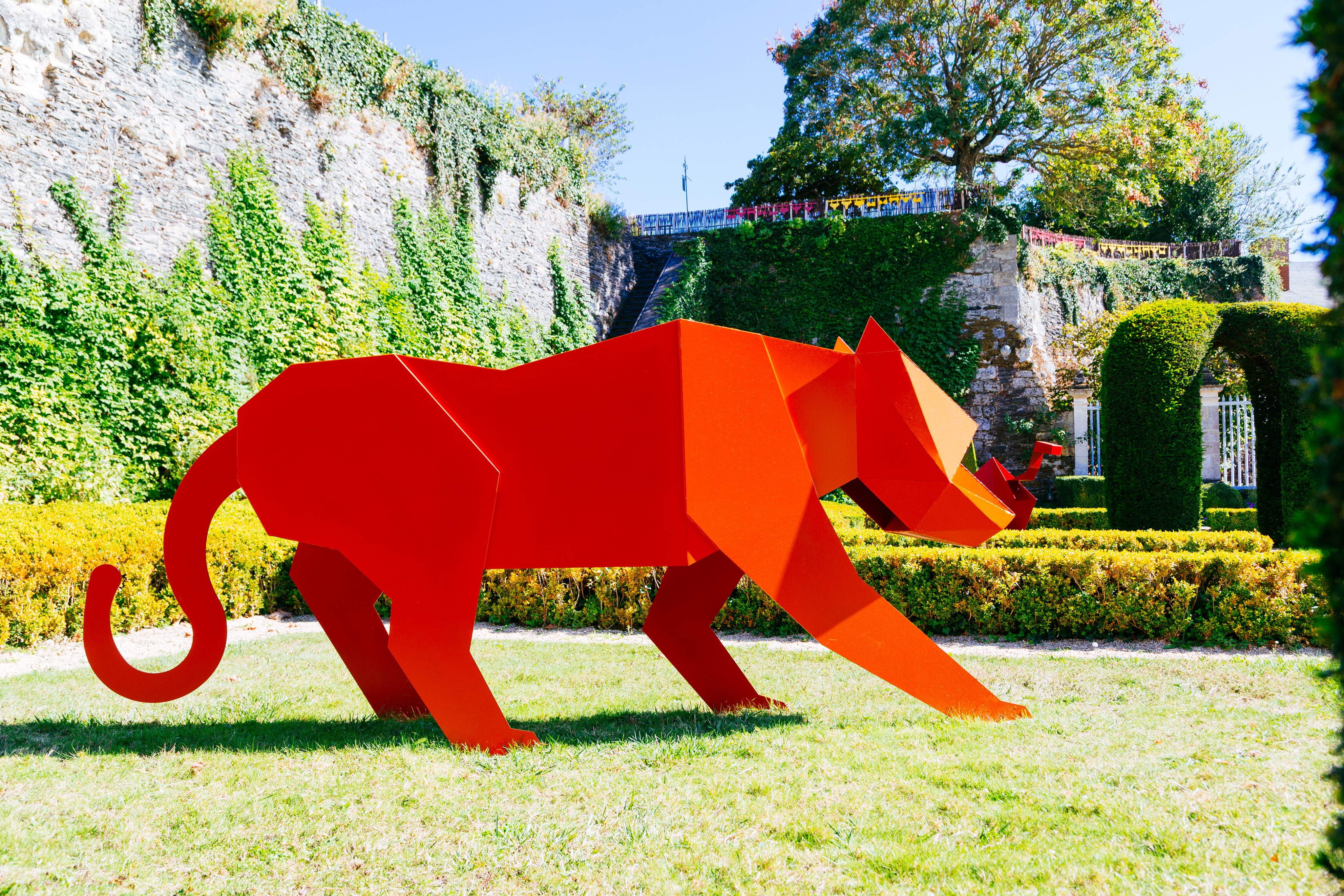 A large orange metal sculpture of a tiger. The statue is in the style of an origami animal. In the background is a groomed garden and a large stone wall.