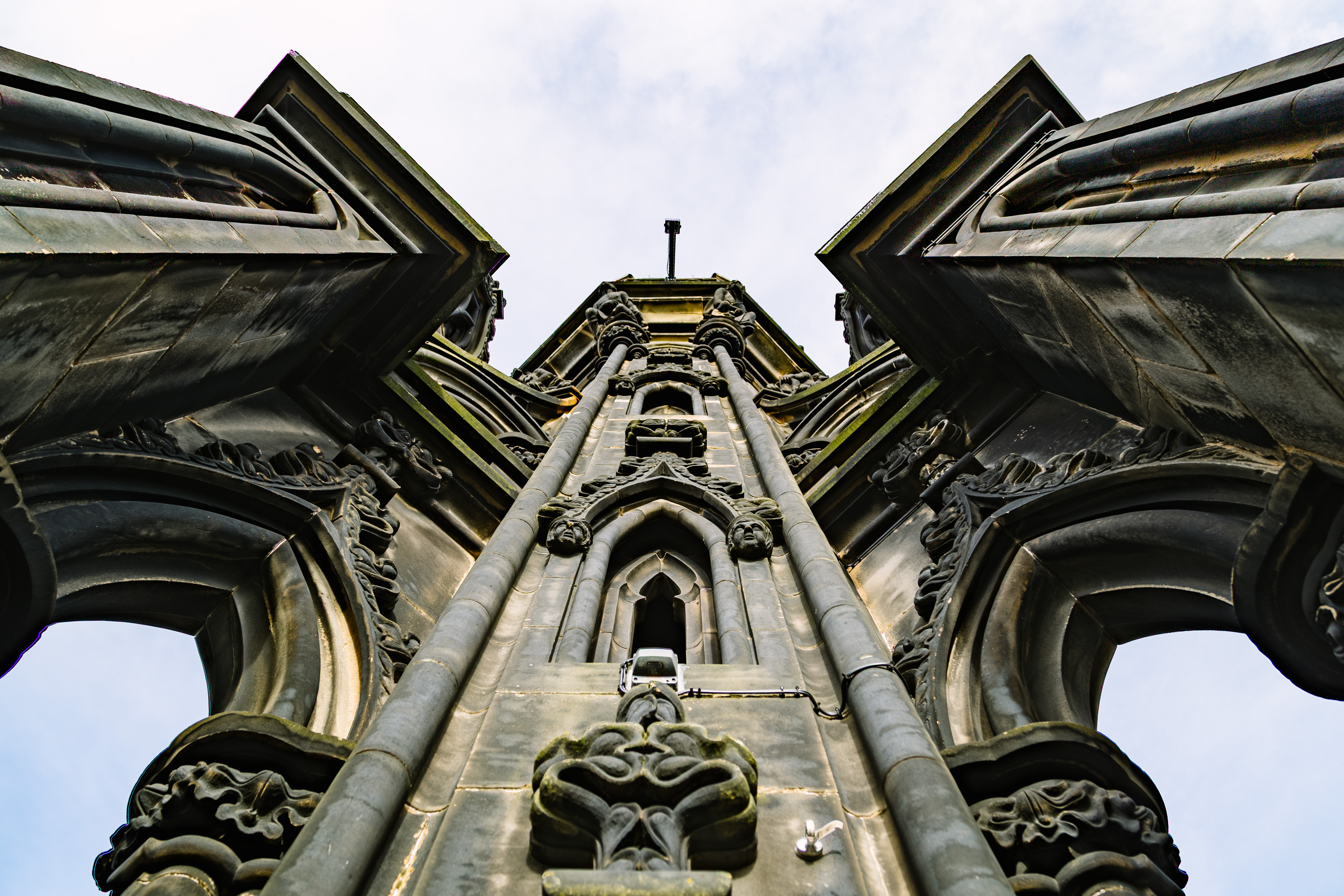A view looking directly up at the Scott Monument in Edinburgh. The view has two arches that are part of flying buttresses on the left and the right. The center is a tower that continues on. The tower is made of stone and is filled with Gothic carvings and adornments: faces, flowers, plants, depictions of animals, etc.