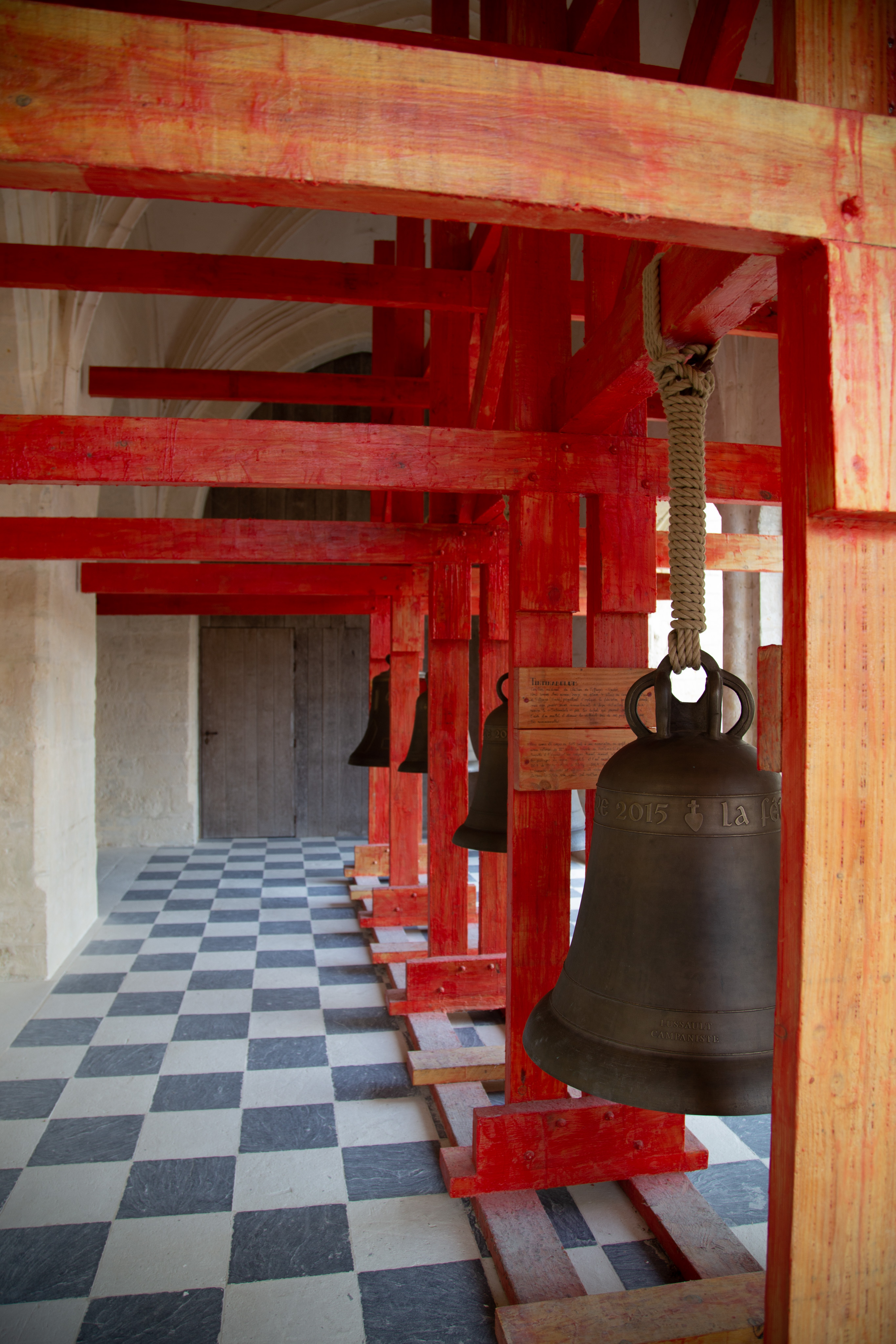 A red, wood frame holds 4 different bells in a row. The bells are each a different size and shape. Each bell is held on to the red frame with rope. The floor is a checkerboard pattern of black and white.