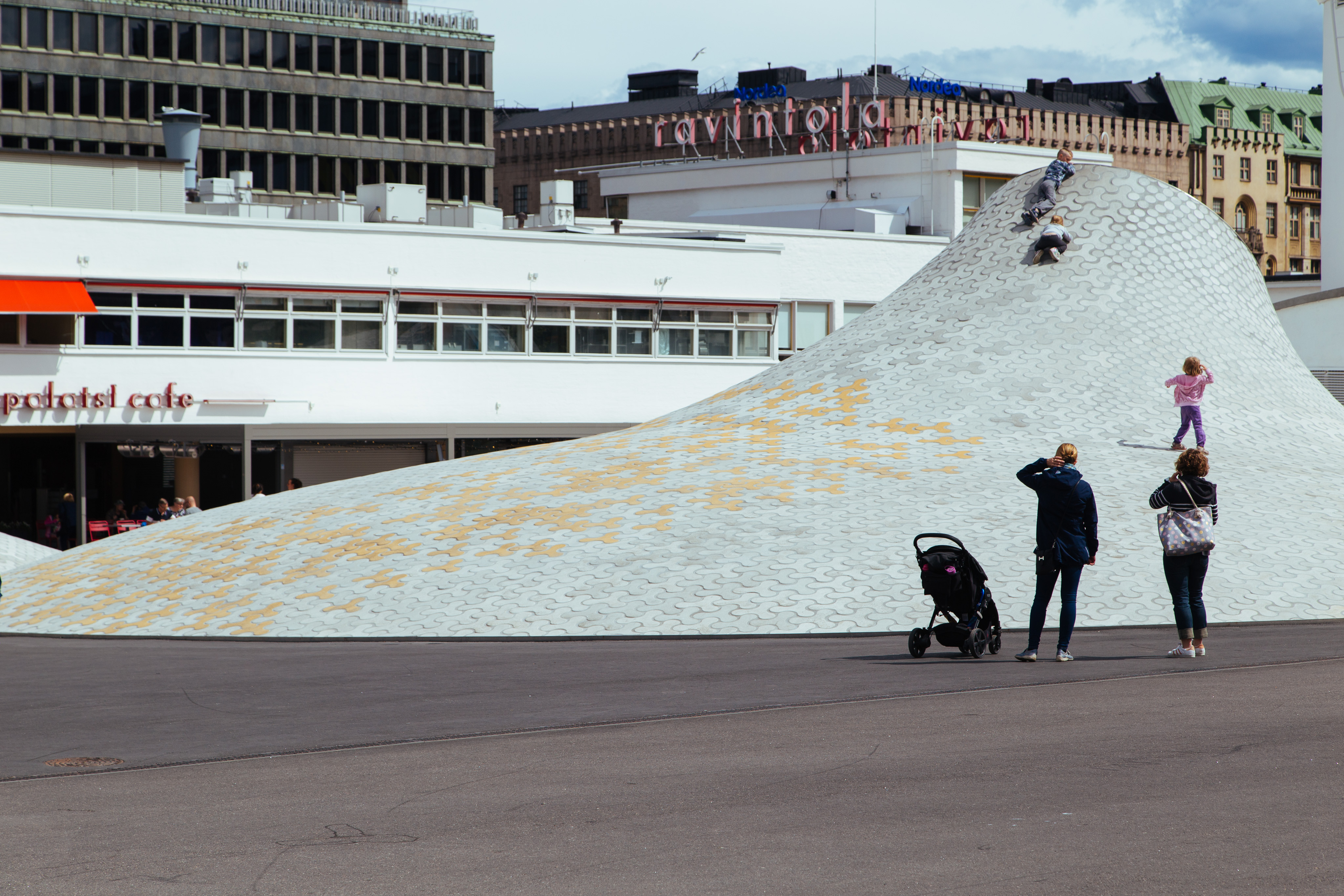 Three children climb up a gently sloping and undulating skylight that pokes out of the ground. Two parents look on.
