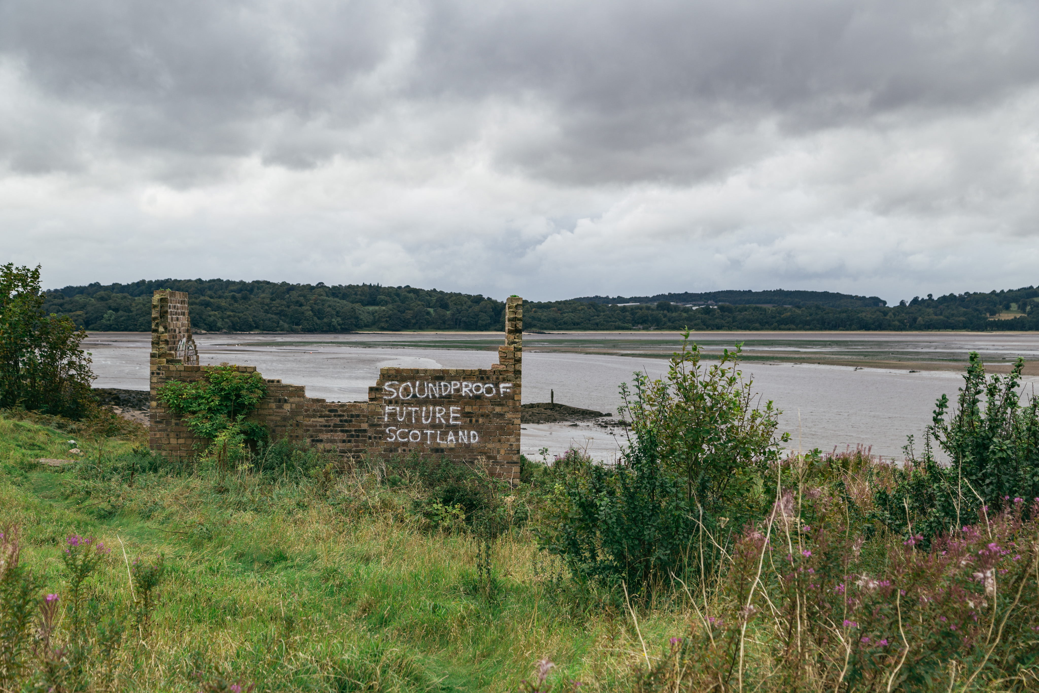 A view from the Island of Cramond. In the midground, there are the ruins of what used to be a brick building. Only three of the four walls are standing (and even those are half height). On the wall closest to the camera is the phrase "Soundproof Future Scotland" in white spray paint. In the background, there is a tidal marsh surrounding the island.