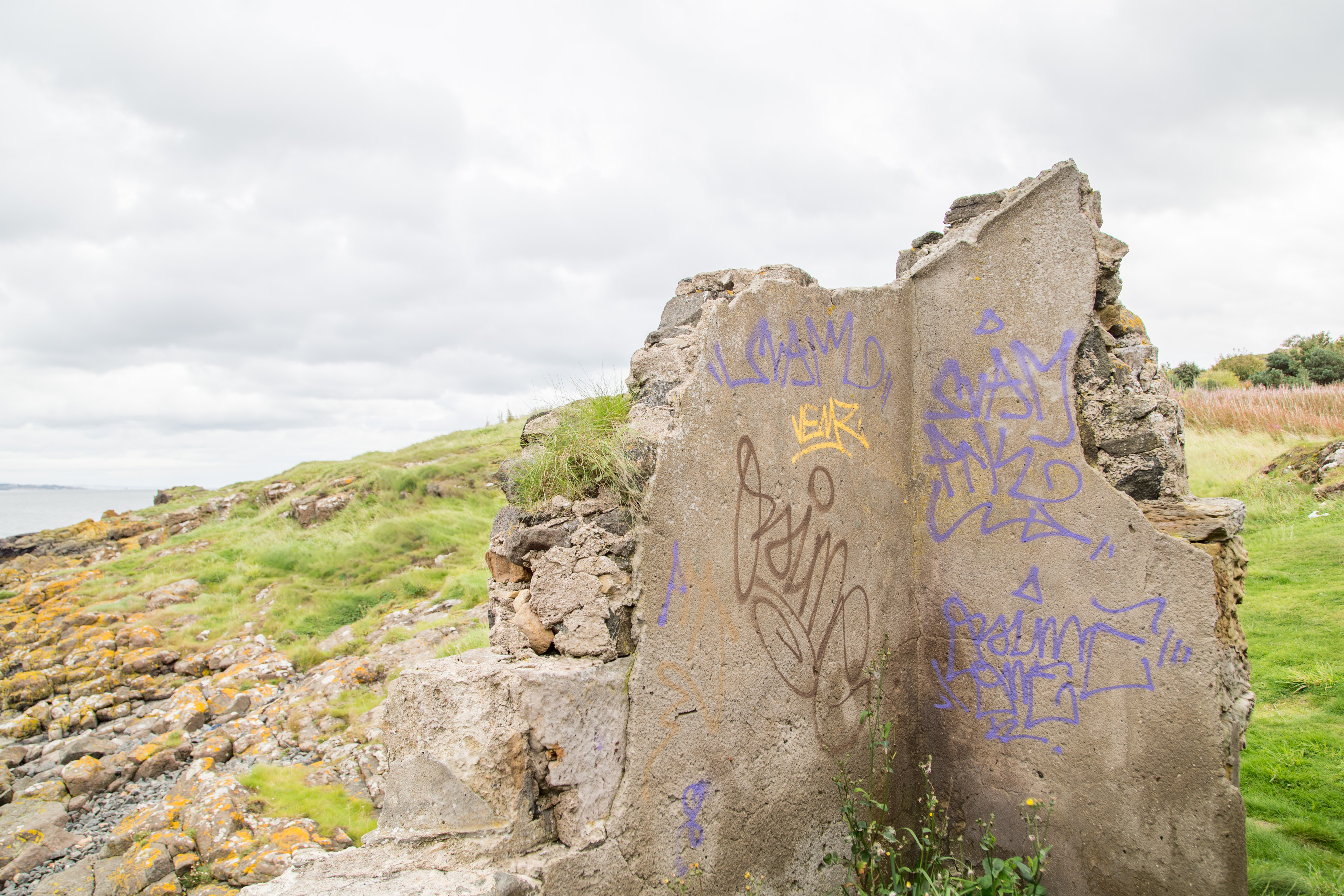 In the center right of the photo are the concrete ruins from the corner of a building that once stood there. There is purple, yellow, and brown spray paint graffiti on both sides of the corner. In the rubble that was once the inside of the walls grass an other plants are growing. In the background is a sloping hill of grass and rock, the Firth of Forth is just barely visible to the left