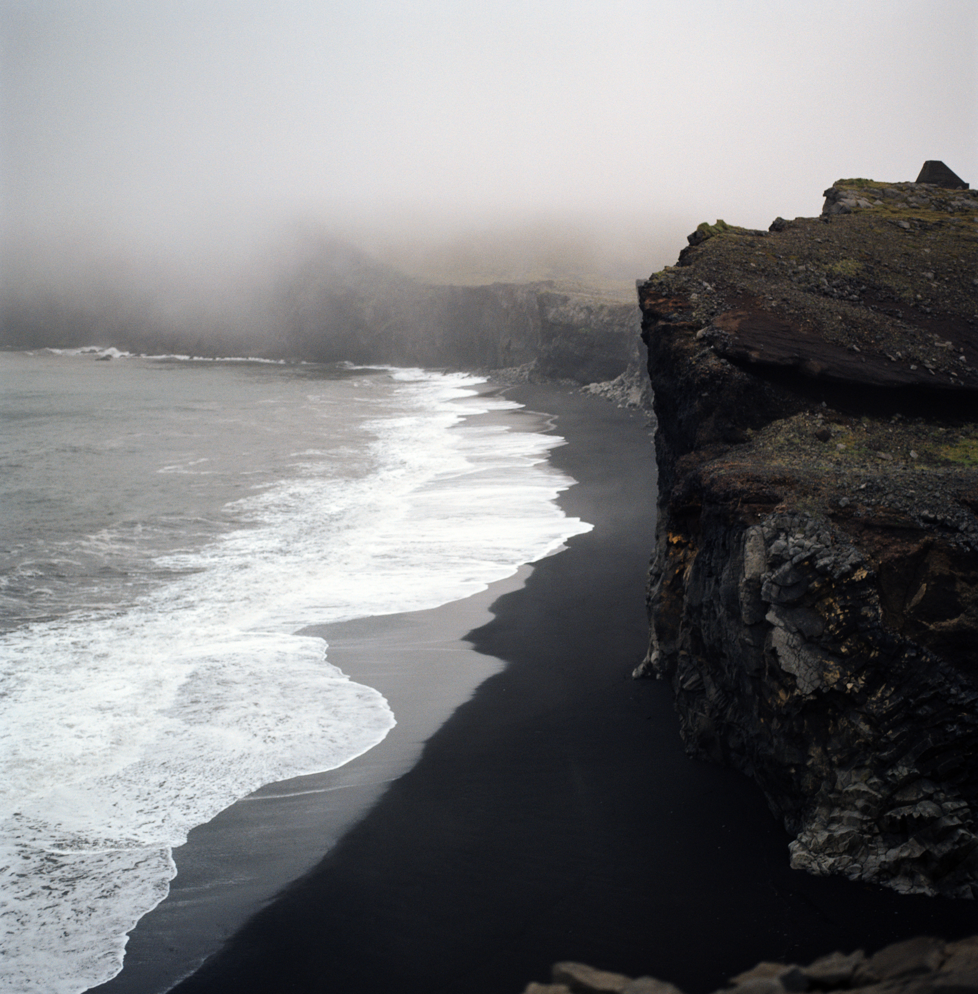 A photo of a black sand beach. There are high cliffs on the right, and there is the white of waves that have broken from the sea on the left.