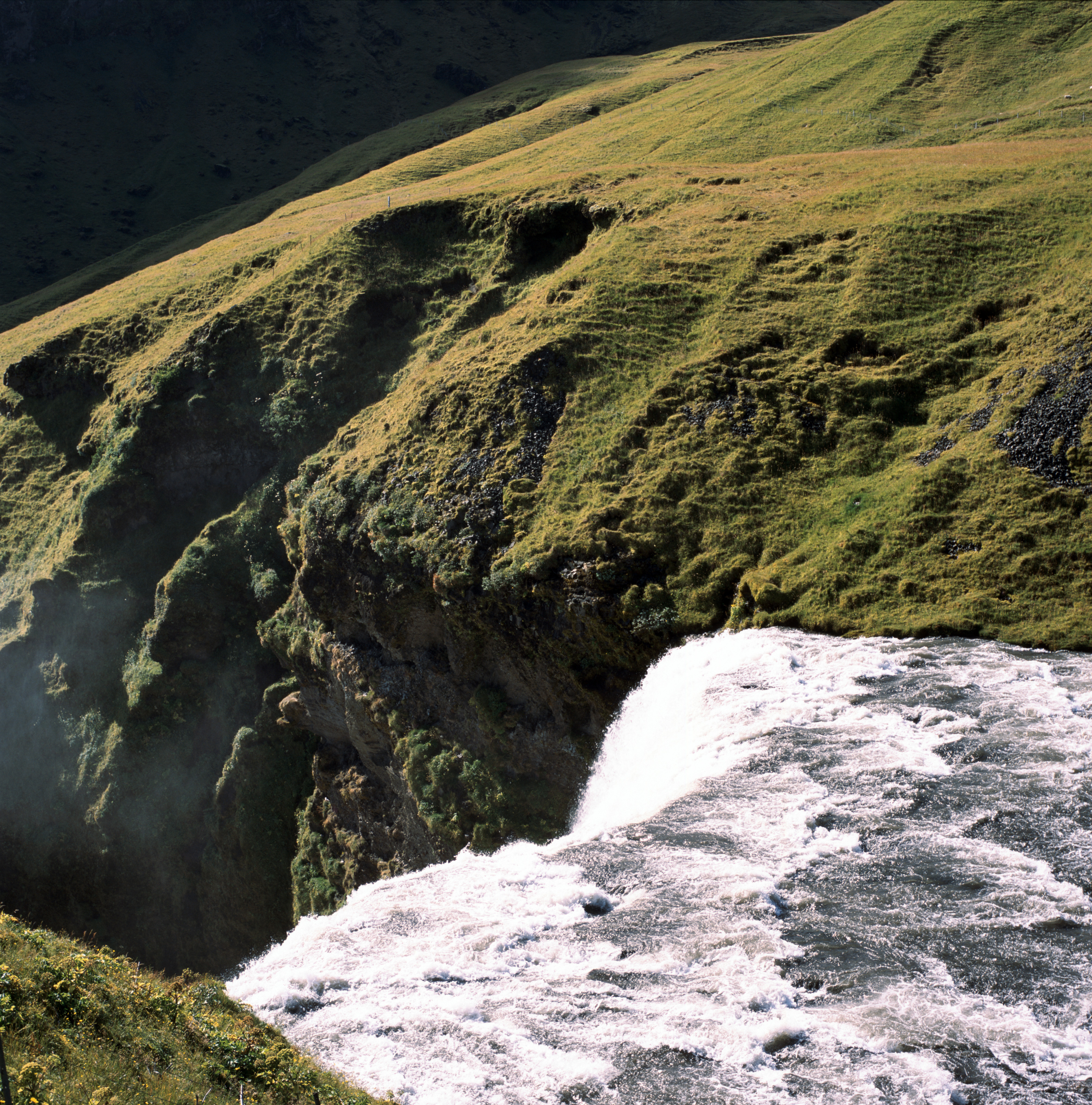 The top of a waterfall from above. The water rushes from the right in the bottom of the frame. It is frozen in time with rocky waves. In the upper part of the frame in the background are rocky cliffs covered in green grass and moss.