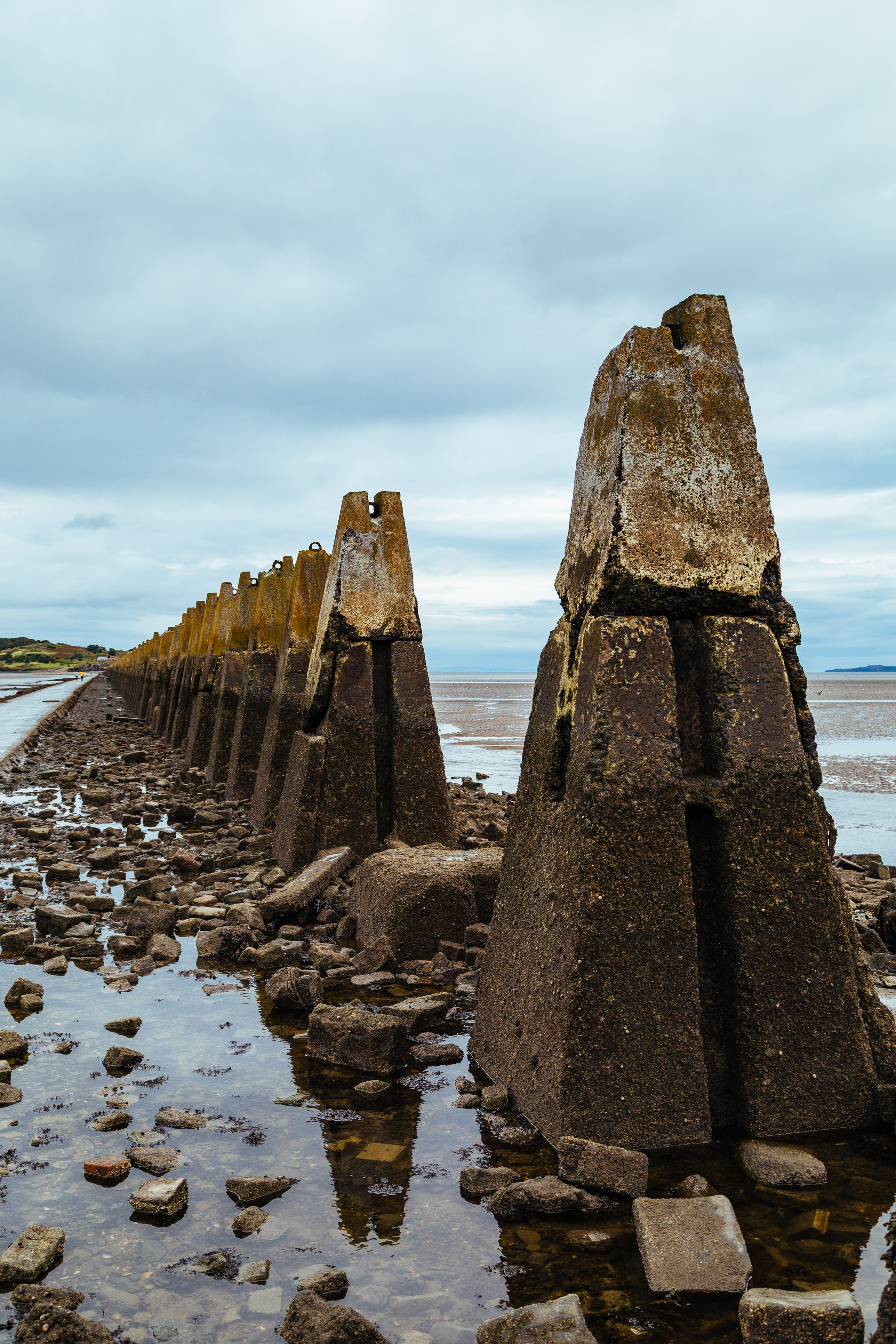 A series of stone pyramids stand in the water along the path to Cramond. The pyramids themselves are concrete and dark on the bottom, but lighter on the top. a path leading to the tidal island is to their left. There are low clouds in the sky in the background.