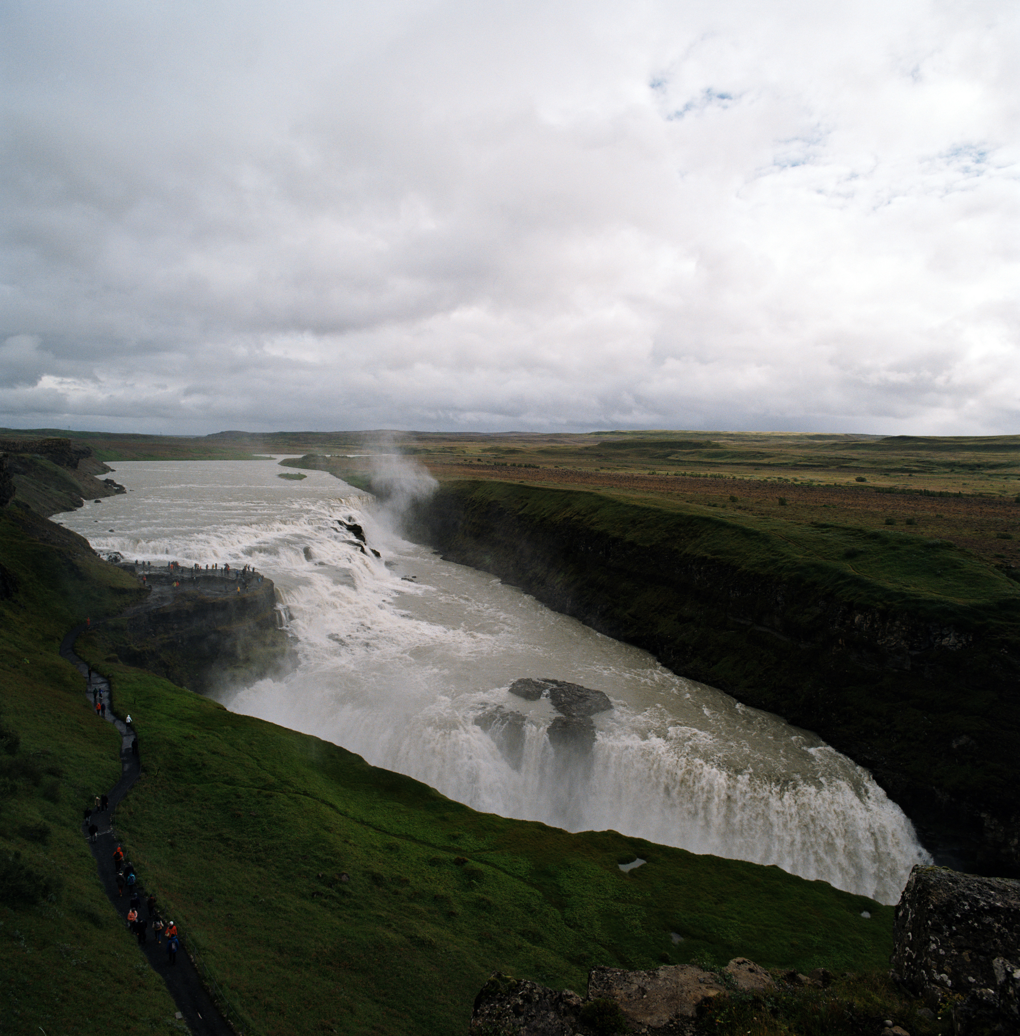A view overlooking Gullfoss in iceland. There are green cliffs on the left and the right, water flowing from the back of the frame towards the falls which disappear behind one of the cliffs. There’s a line of people walking along a black path to the left