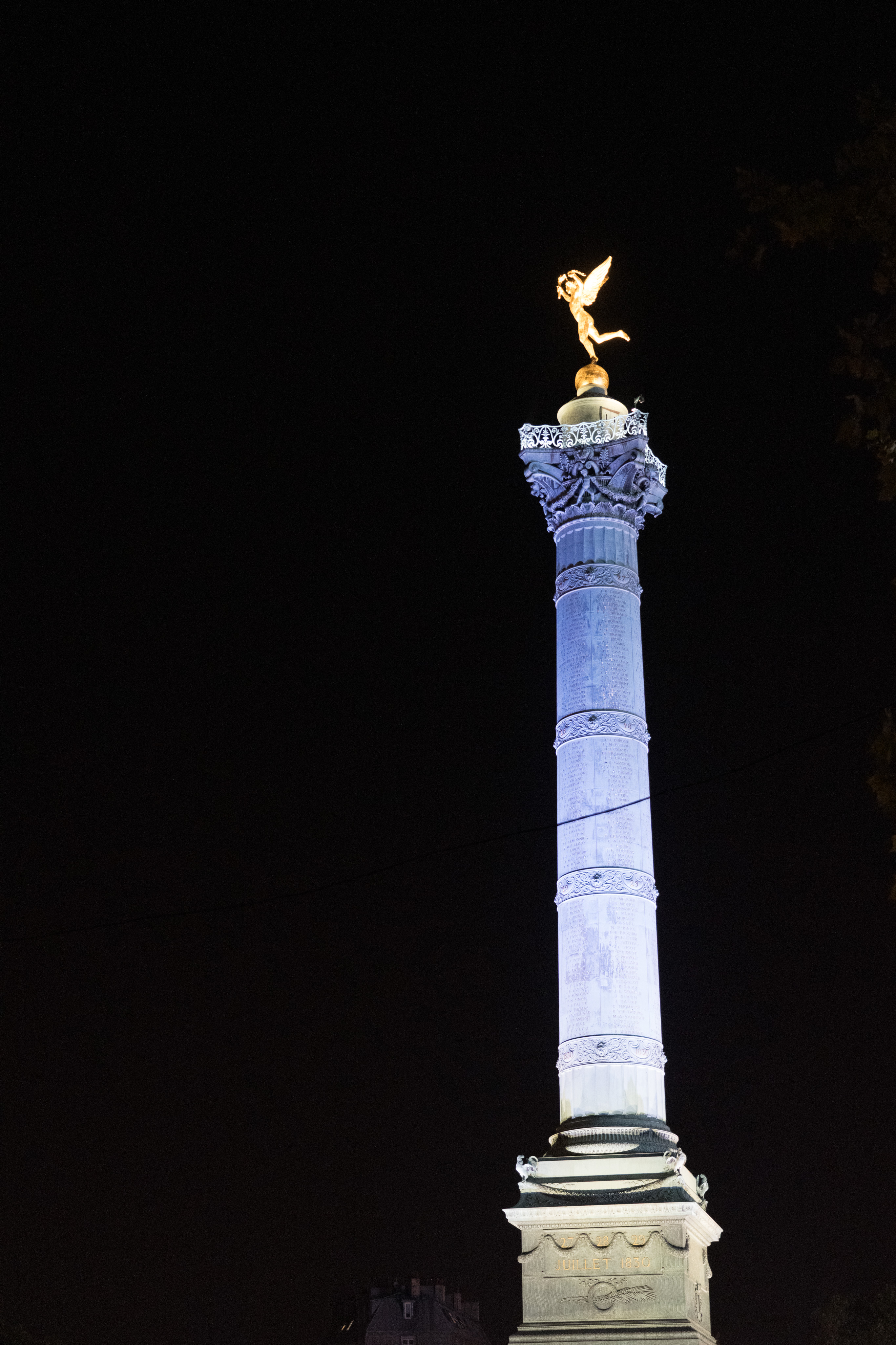 A view of the Colonne de Juillet (english: The July column). A single, grey Corinthian column stands alone on a pedestal. On top of the column is a gold statue of an angel with wings. The angel is brandishing a torch and broken chains. The background is all black.